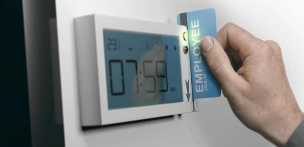 Eine Person schiebt ihre Mitarbeiterkarte in ein Gerät für die elektronische Zeiterfassung, auf dessen Display die Uhrzeit 07:59 AM steht.